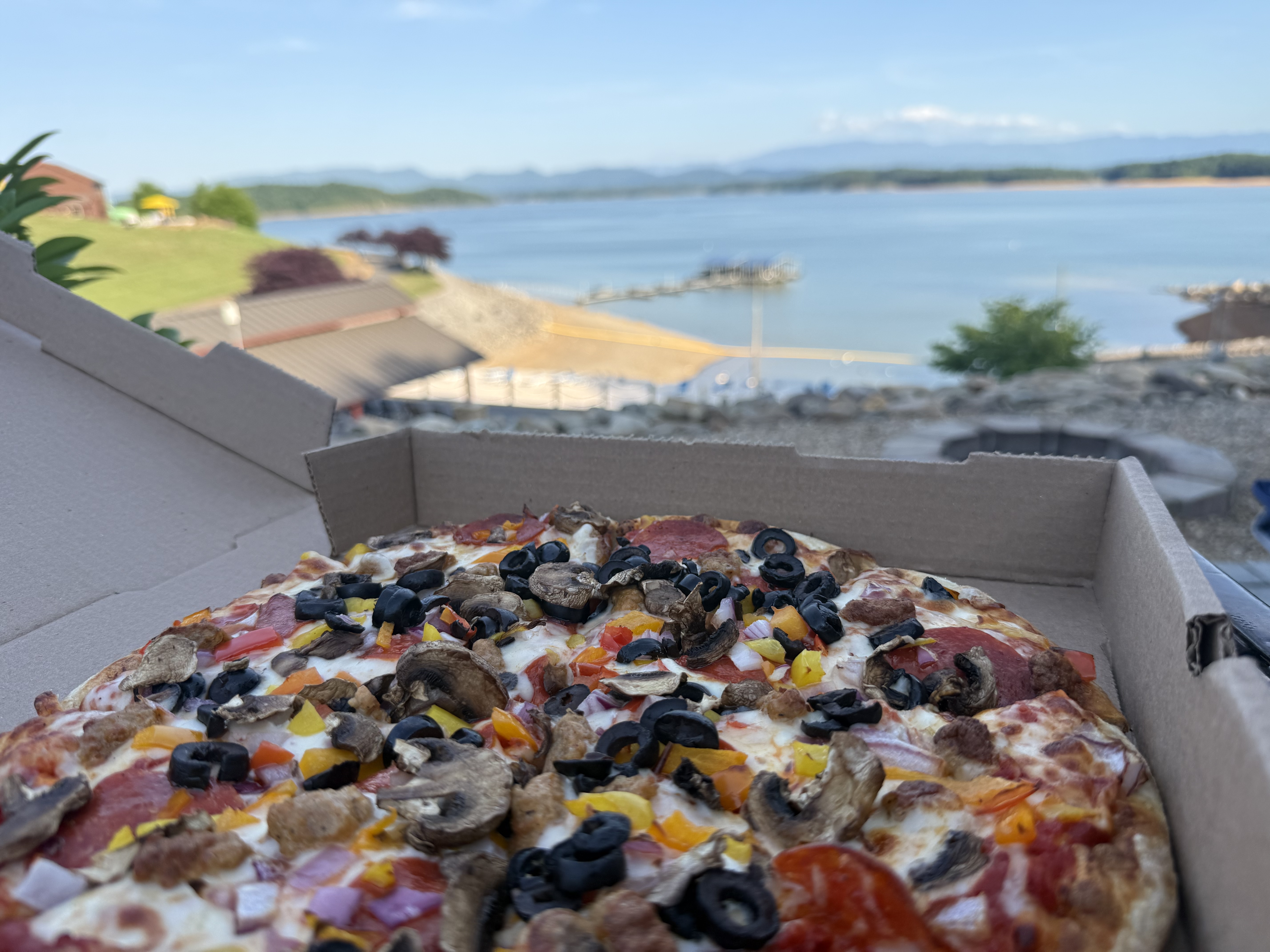 A loaded supreme pizza in an open box at an RV site, with Douglas Lake and the mountains blurred behind it.